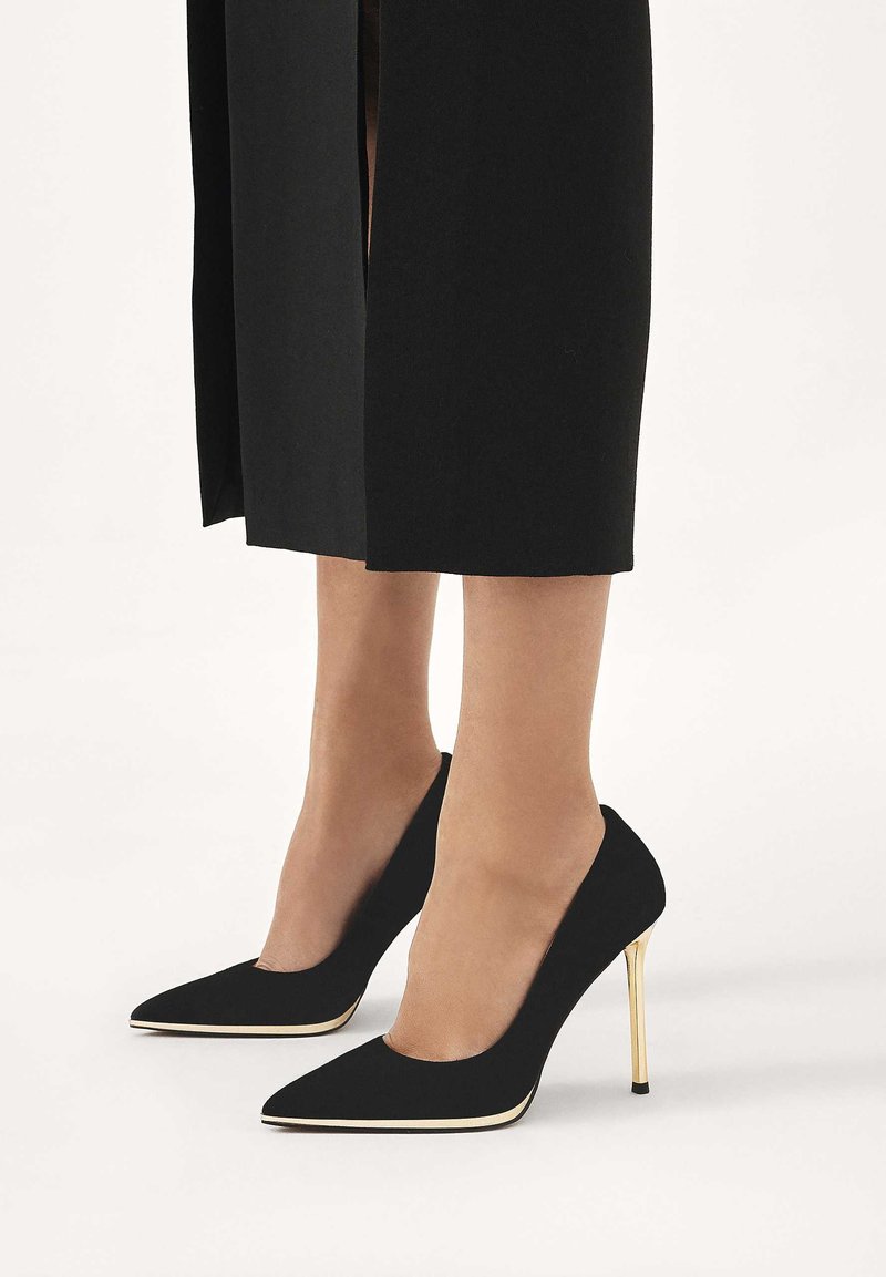 Mustad seemisnahast kontsakingad terava ninaosaga, mille tallal on kuldne detail, taustal tavaline taust ja must riideese.