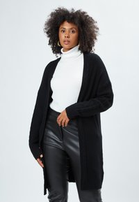 Cardigan nero con design a trecce, maniche lunghe e apertura frontale. Indossato sopra un dolcevita bianco, abbinato a pantaloni in pelle nera.