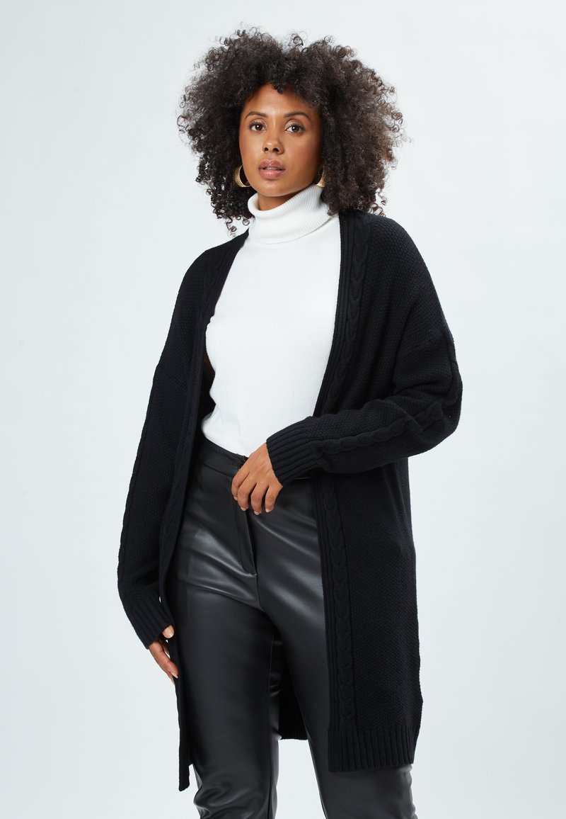Cardigan nero con design a trecce, maniche lunghe e apertura frontale. Indossato sopra un dolcevita bianco, abbinato a pantaloni in pelle nera.