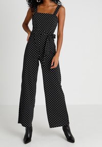 Svart prickig jumpsuit med fyrkantig halsringning, vid ben-design och knytbälte i midjan. Tillverkad av lättviktsmaterial, i kombination med klackboots.