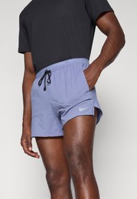 Helllila Laufshorts mit perforierter Textur, schwarzem Zugband am Bund und reflektierendem Nike-Logo auf der Vorderseite. Getragen mit einem schwarzen Oberteil.