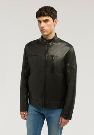 Joven con chaqueta de cuero negra con cremallera y cuello con botones a presión, y pantalones vaqueros azules, de pie frente a un fondo claro y liso.