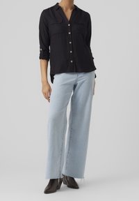 Camicia nera con bottoni, due tasche sul petto e maniche arrotolate, abbinata a jeans a gamba larga azzurri e stivali neri alla caviglia.