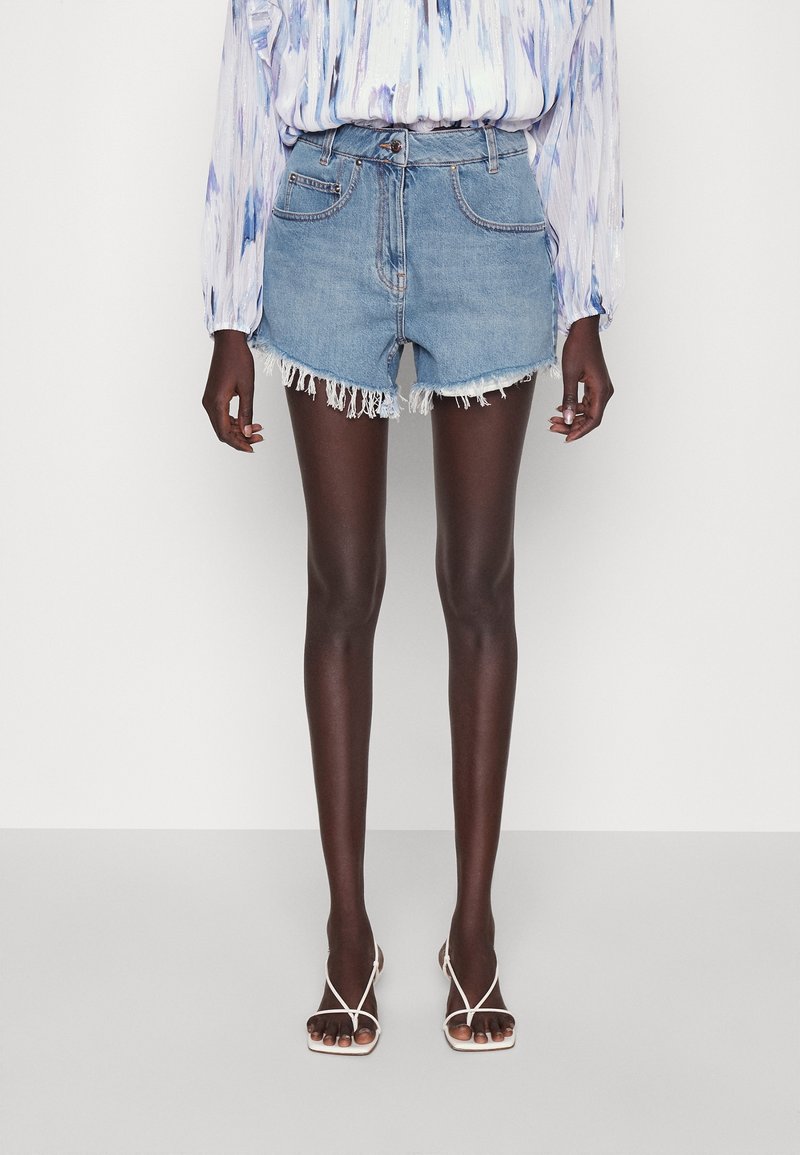 Femme portant un chemisier tie-dye bleu clair, un short en denim déchiré taille haute et des sandales blanches à talons avec des lanières, debout sur un sol blanc.