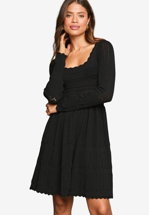 Rochie tricotată neagră cu decolteu scallop, mâneci lungi cu accente texturate și fustă nivelată cu o siluetă evazată.