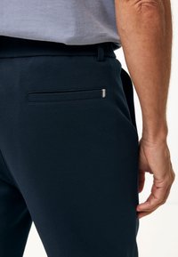 Pantaloni navy con una texture liscia e raffinata, una singola tasca posterior e un'apertura su misura, oltre a un sottile dettaglio argentato sulla tasca.