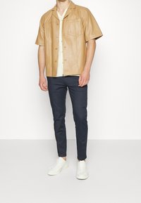 Man i en beige kortärmad läderskjorta över en krämfärgad skjorta, marinblå slim-fit byxor och vita sneakers, som står mot en neutral bakgrund.