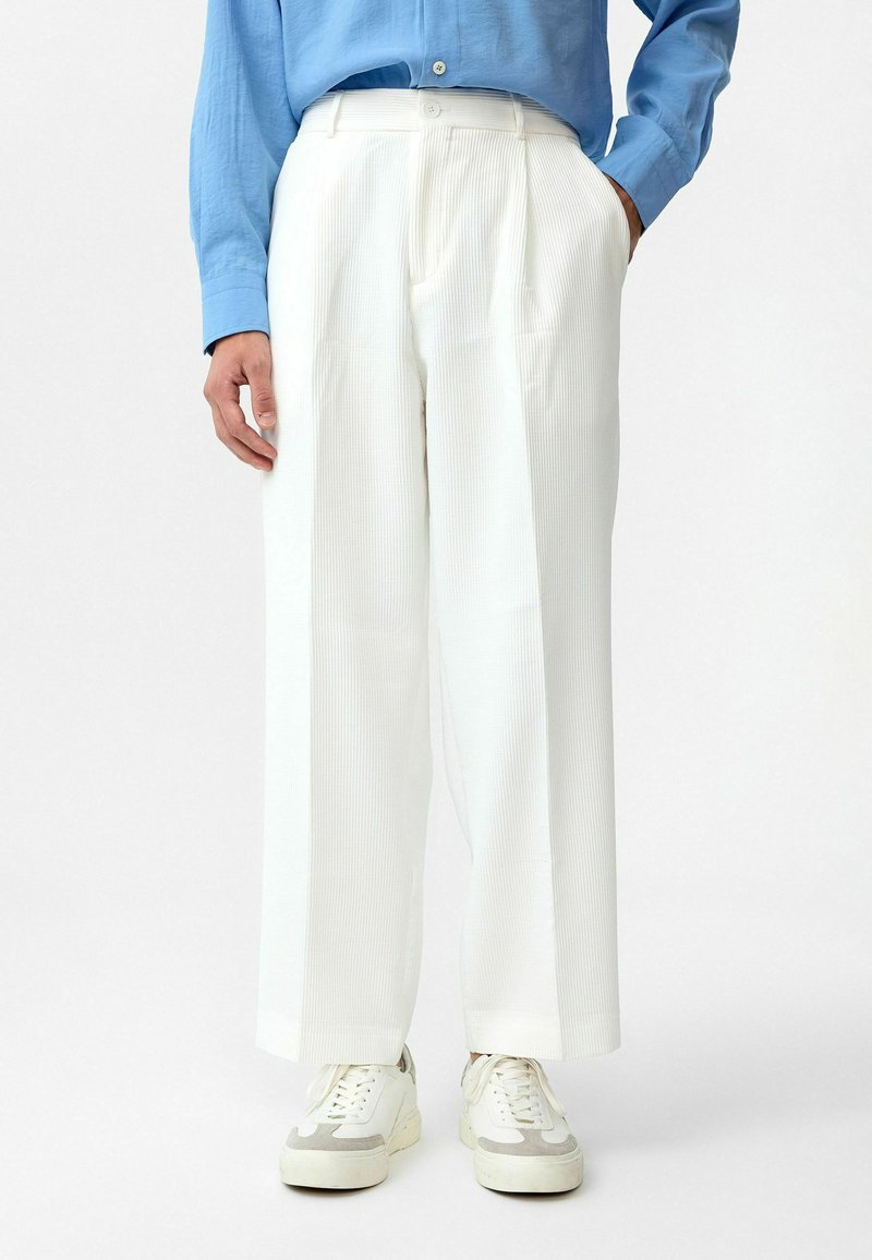 Witte, geplooide broek met een wijdlopende pasvorm; textuurstof; gecombineerd met lichtgekleurde sneakers; gedragen met een blauwe lange mouw shirt.