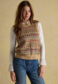 Kudumine beež vest, millel on mitmevärvilised lille- ja geomeetrilised mustrid, kihiline valge bluus, millel on volangidega varrukad, koos siniste teksadega.