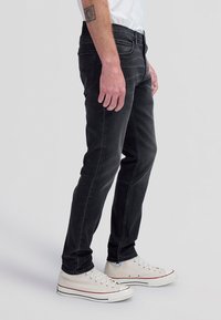 Mann trägt schlanke schwarze Jeans, weiße Sneakers und ein weißes Hemd, steht mit entspannten Armen seitlich am Körper vor grauem Hintergrund.