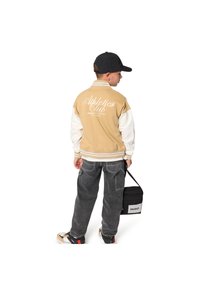 Μπεζ και λευκό μπλουζάκι μπέιζμπολ με το κείμενο «Athletics Club», μαύρο καπέλο, γκρι τζιν παντελόνι και μαύρη τσάντα με λευκό λογότυπο. Τα αθλητικά παπούτσια έχουν απαλά χρώματα.