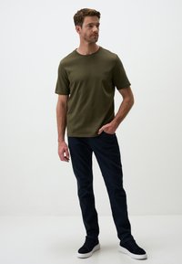T-shirt verde oliva a maniche corte con colletto a girocollo, abbinata a pantaloni slim-fit blu scuro e sneakers nere con suola bianca.