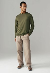 Olivengrünes Langarmhemd kombiniert mit hellbeigen Hosen. Das Modell trägt braune und graue Sneakers und steht vor einem neutralen Hintergrund.