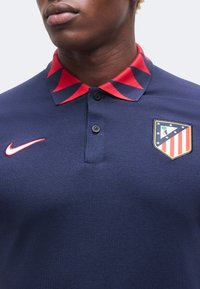 Homme portant un polo bleu marine avec un col géométrique rouge, un patch logo Atletico Madrid et un swoosh rouge Nike sur la poitrine.
