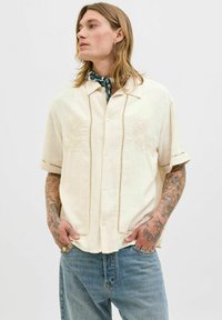 Camisa de manga corta color crema con diseños y acentos florales bordados, que presenta un cierre de botones y un corte relajado.