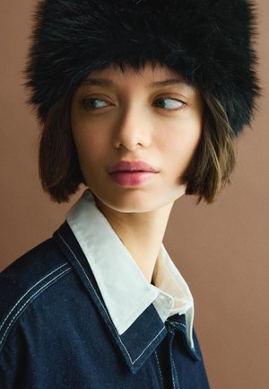 Chapeau en fourrure noire avec une texture douce, associé à une veste en denim foncé avec un col blanc. L'arrière-plan est d'une couleur chaude et neutre.