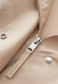 Tessuto beige con una consistenza liscia, dotato di una cerniera argentata e di hardware con bottone a scatto. L'etichetta recita "OPUS" vicino al tirante della cerniera.