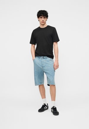 Jeune homme debout, portant un t-shirt noir, un short en jean bleu clair, des chaussettes blanches et des baskets noires sur un fond blanc uni.
