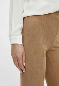 Brązowe spodnie o teksturowanej powierzchni z pionowymi szwami, zestawione z białą bluzką, prezentujące zbliżenie na tkaninę i ułożenie dłoni.