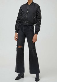 Veste bomber noire avec fermeture éclair, dotée de poignets et d'un ourlet côtelés. Associée à un jean noir taille haute évasé avec des découpes aux genoux effilochées.