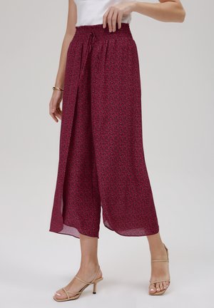 Vrouw draagt wijde, enkelhoge bordeauxrode broek met een klein geometrisch patroon en een elastische tailleband, gecombineerd met beige sandalen met hak.