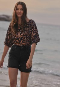 Model nosi tamnu majicu s uzorkom svjetlosmeđi listova i crne traper kratke hlače visokog struka s preklopom u blizini plaže.