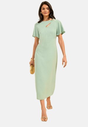 Frau trägt ein hellgrünes Midikleid mit Flatterärmeln und kleinem Ausschnitt an der Brust, hält eine goldene Clutch und trägt goldene Sandalen.