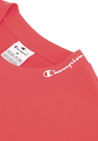 Camiseta de algodón roja con cuello redondo. Presenta un logo blanco de "Champion" en el cuello. Etiqueta de tamaño visible en la tela.