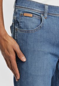 Vaqueros de mezclilla azul con corte ajustado, que cuentan con un bolsillo frontal y una etiqueta de marca de cuero cerca de la cinturilla. Textura suave y detalle de costura clásico.