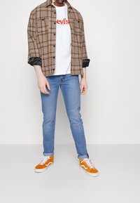 Camisa de cuadros marrón de botones sobre una camiseta blanca con logotipo rojo, jeans de mezclilla azul y zapatillas naranjas con detalles blancos.