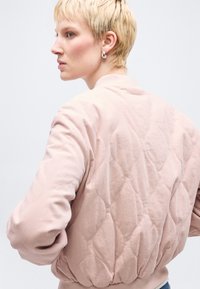 Γυναίκα με κοντά ξανθά μαλλιά, που φορά ασημένια σκουλαρίκια κρίκους και ένα ανοιχτό ροζ παλτό με καπιτονέ σχέδιο, κοιτάζει προς τα αριστερά πάνω σε λευκό φόντο.