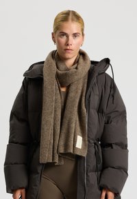 Brun uld tørklæde med en blød tekstur, draperet omkring halsen. Vedhæftet label med teksten "rethink" på kanten. Båret over en sort pufferjakke.