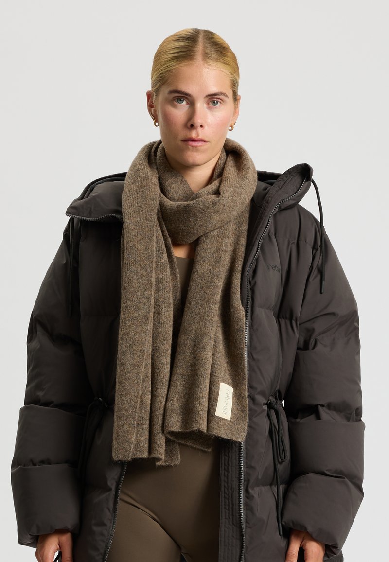 Brun uld tørklæde med en blød tekstur, draperet omkring halsen. Vedhæftet label med teksten "rethink" på kanten. Båret over en sort pufferjakke.