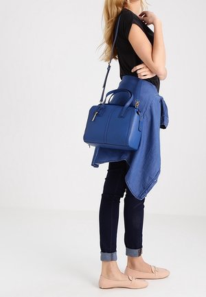 Femme portant un haut noir, un jean foncé retroussé, une chemise bleue nouée à la taille, tenant un sac à main bleu avec bandoulière, debout sur un fond uni.