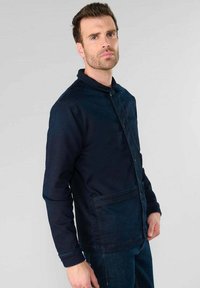 Navyblauwe spijkerjacket met opstaande kraag, knopen aan de voorkant en een grote zak. Kenmerkt zich door contrasterende stiksels en een relaxte pasvorm.