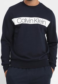 Námořnická mikina vyrobená z měkké tkaniny, s bílým horizontálním pruhem se středovým nápisem "Calvin Klein". Hribované manžety a lem.