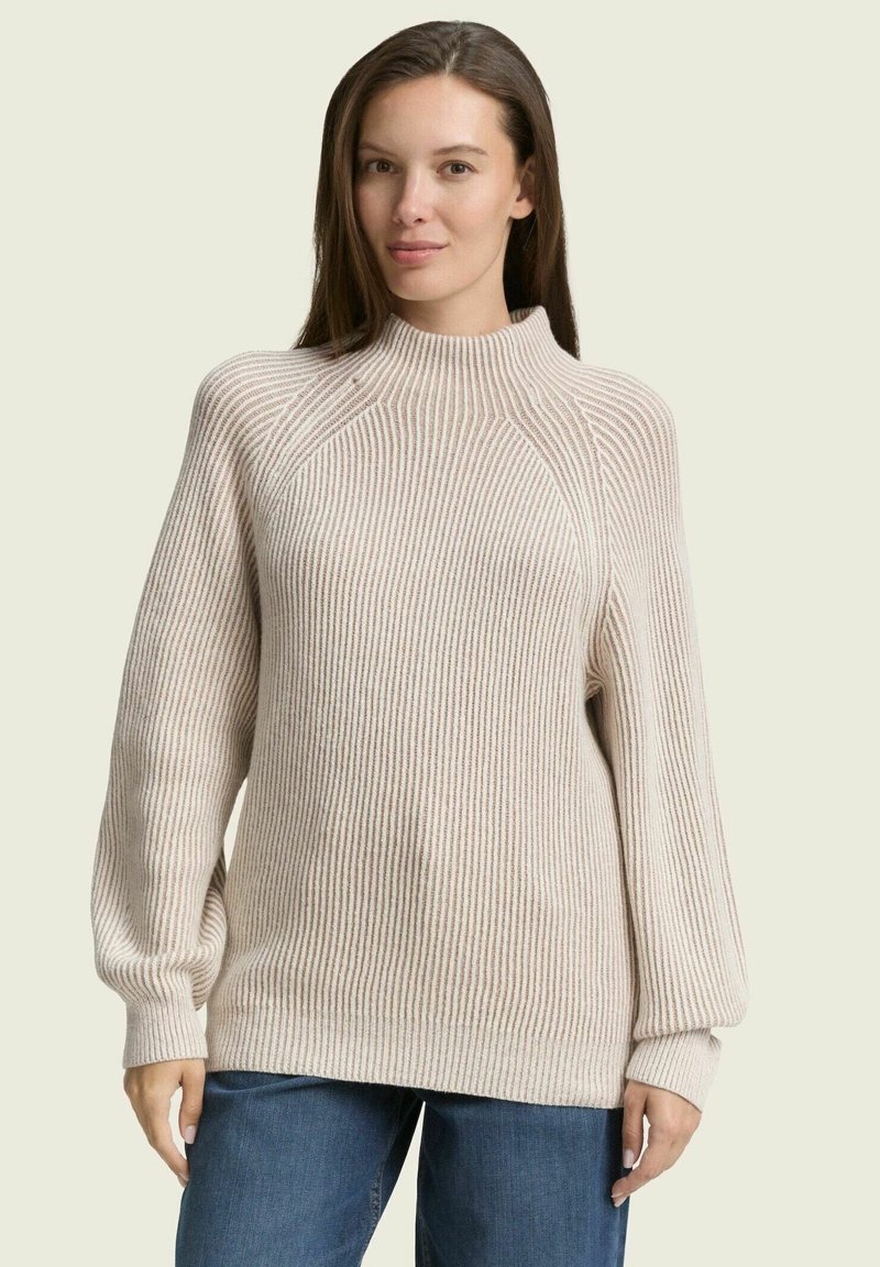 Pull en maille beige avec un col montant, des manches longues ballon et une coupe décontractée, associé à un jean en denim bleu.