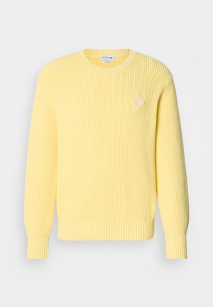 Kollane soonikkoes kampsun ümarate kaelusega, pikkade varrukatega ja rinnal rombikujuline logo. Pehme tekstuur ja keha jälgiv lõige.