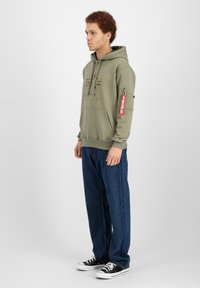Felpa con cappuccio verde oliva con logo frontale, dotata di una tasca laterale e un'etichetta rossa. Abbinata a jeans blu a taglio largo e sneakers nere.