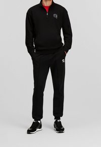 Sudadera negra con cremallera y cuello alto, adornada con un pequeño logo blanco, combinada con pantalones negros ajustados con detalles en blanco y zapatillas negras.