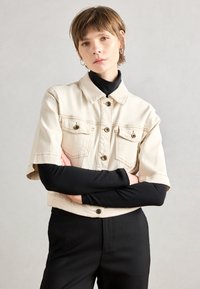 Klippt, beige denimjacka med korta ärmar, två bröstfickor och svarta knappar. Bärs över en åtsittande svart polo.