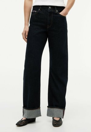 Person iført mørkeblå jeans med vide ben og op rullede kanter, hvid striktrøje og sorte vævede flade sko.