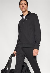 Μαύρο τζάκετ με φερμουάρ από απαλό ύφασμα, με ψεύτικο γιακά, μπροστινές τσέπες και κεντημένο λογότυπο. Στυλιζαρισμένο με παντελόνι και τσάντα στο ίδιο σχέδιο.