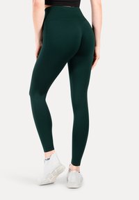 Donkergroene high-waisted leggings van rekbaar materiaal, met een gladde textuur en een strakke pasvorm. Gecombineerd met witte sportschoenen.