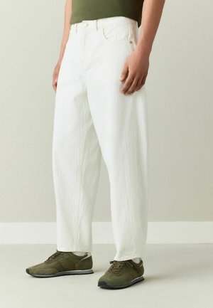 Person wearing loose white pants, olive green sneakers, and an olive green shirt, standing against a plain light background.