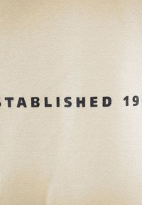 Tessuto beige con un bold testo nero che recita "FONDATO NEL 19" posizionato centralmente. Texture liscia con un sottile motivo a maglia.