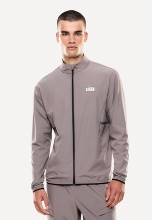 Giovane uomo che indossa una giacca EA7 taupe con cerniera e pantaloni coordinati, in piedi con un'espressione neutra su sfondo bianco.