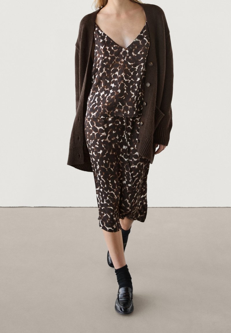 Femme portant une robe imprimé léopard marron foncé avec un décolleté en V profond, un cardigan marron foncé, des chaussettes noires et des mocassins noirs marchant vers l'avant.