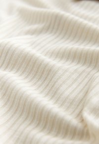 cremefarbener, gerippter Strickstoff mit weichen Falten und feiner Textur, der dezente Schatten und Lichtreflexe zeigt.