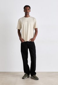 Beige t-skjorte med korte ermer og brodert logo, kombinert med svarte bukser med rette ben og olivengrønne sneakers med grå såler.